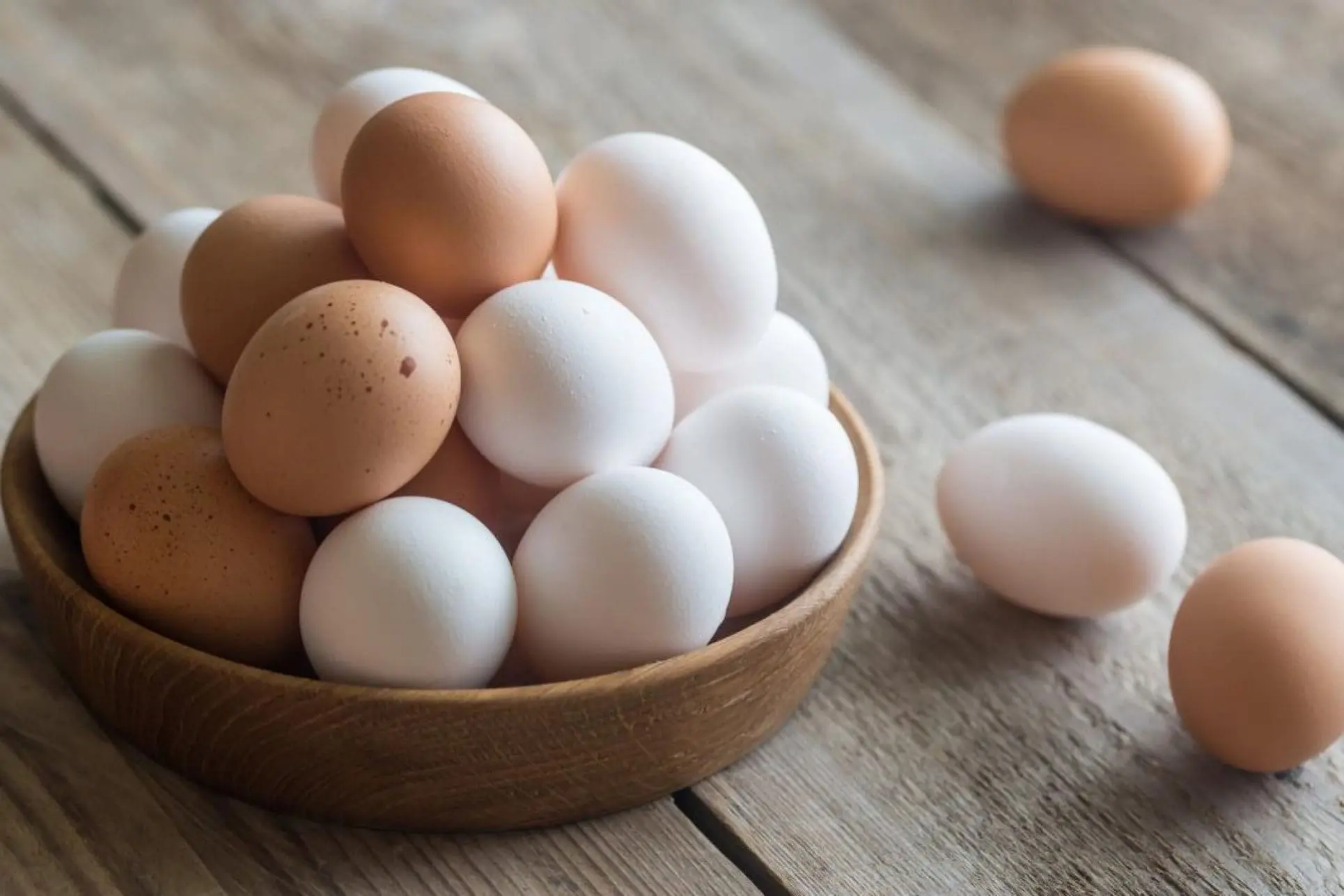 تخم مرغ هایی به رنگ سفید و قهوه ای که در ظرف چوبی قرار دارد و در اطراف ظرف نیز تخم مرغ وجود دارد