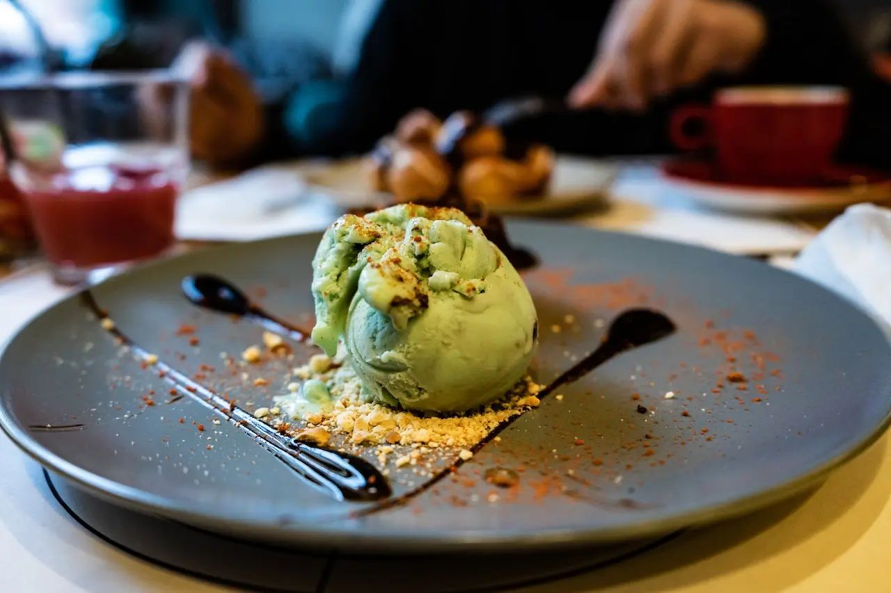 بستنی پروتئینی سبز که در بشقاب مشکی قرار دارد و سس بستنی شکلاتی هم در اطراف آن ریخته شده است